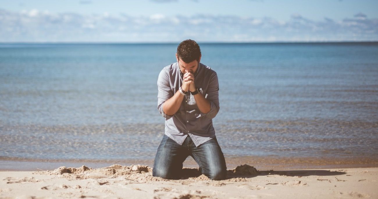 man kneeling on a beach praying with his head bowed